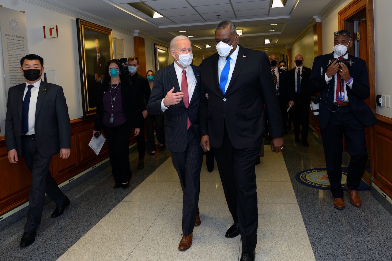 Two men dressed in business suits walk down a hall and talk followed by other people.