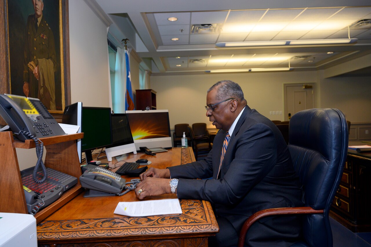 Secretary of Defense Lloyd J. Austin III speaks while sitting at a desk with a phone on it.