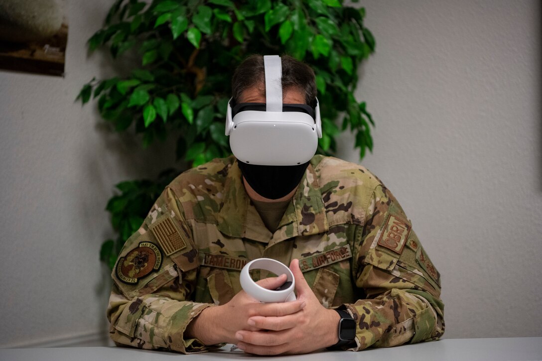 A man in uniform sits at a table weating a virtual reality headset while holding a virtual reality controller.