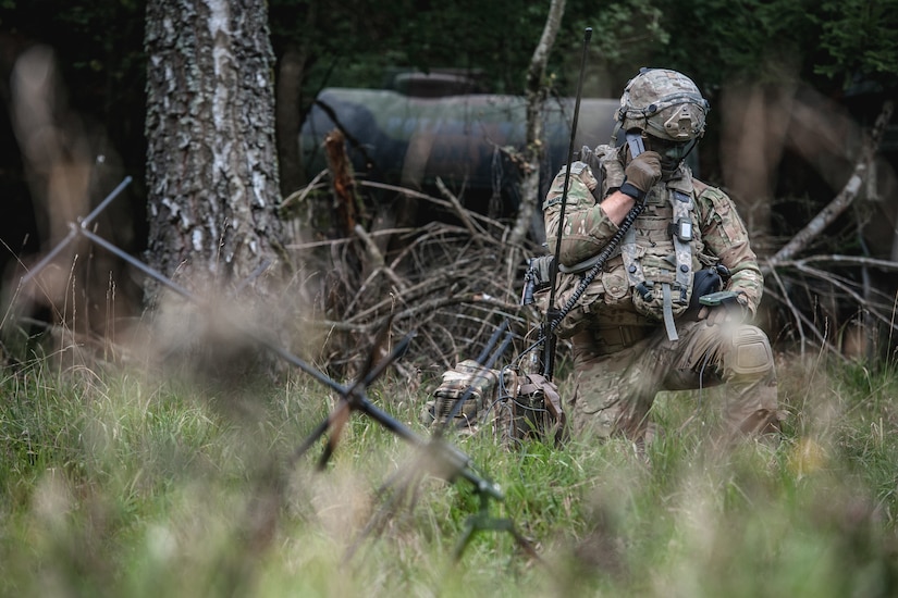 A soldier in a wooded area talks on a radio.