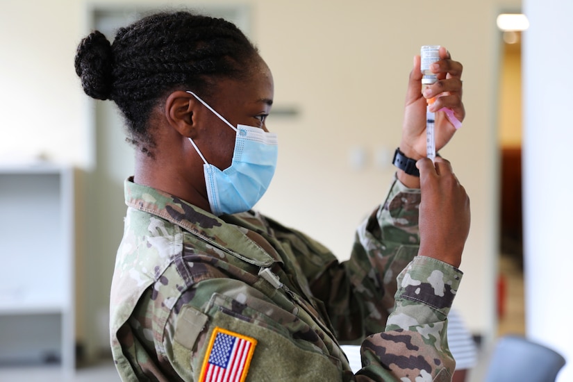 A soldier wearing a mask holds a syringe with the needle inserted into a small bottle.