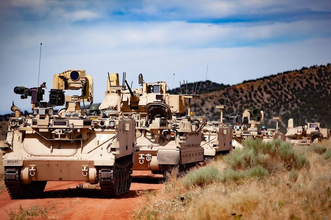 Tactical vehicles in single-line formation travel down dirt road,