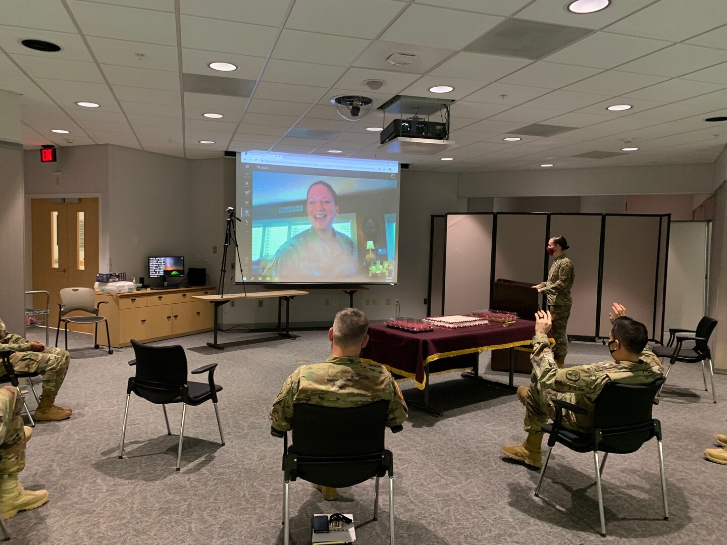 Soldiers sitting in chairs in a classroom while a soldier is talking virtually and shown on a movie screen.