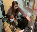 A soldier in uniform is sitting while talking on the phone. A civilian stands behind her in the office pointing at a computer screen.