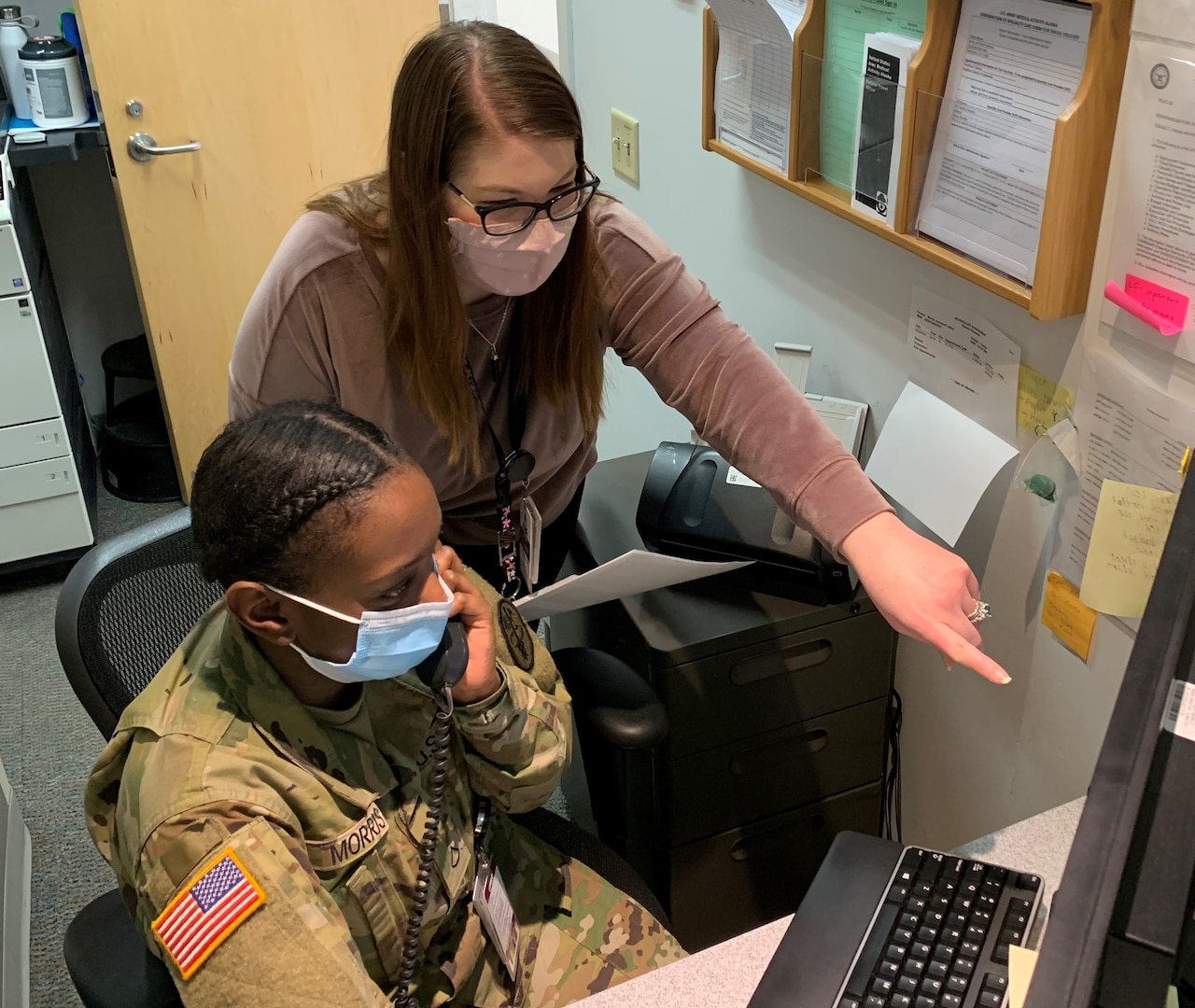 A soldier in uniform is sitting while talking on the phone. A civilian stands behind her in the office pointing at a computer screen.