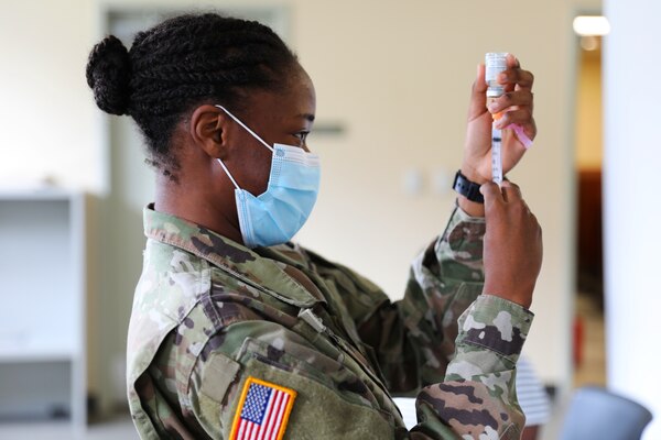 A soldier wearing a mask holds a syringe with the needle inserted into a small bottle.