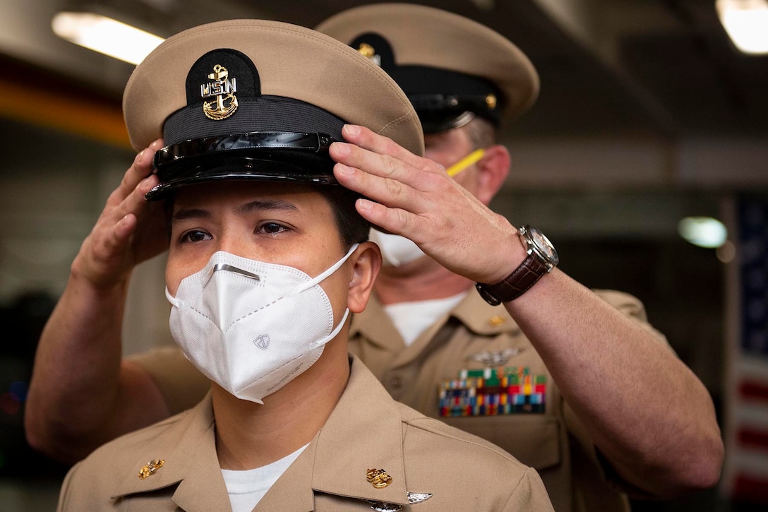 A Navy cover is placed on the head of a sailor by another sailor standing behind her.