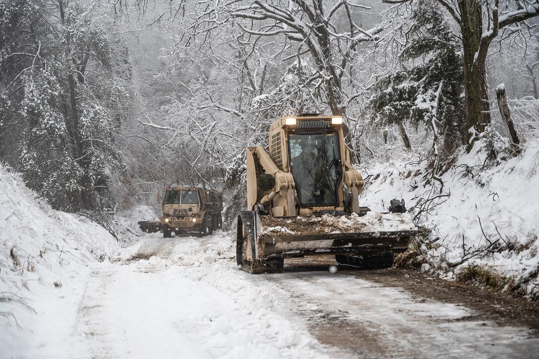 A bulldozer and a plow clear debris from a snow-covered road.
