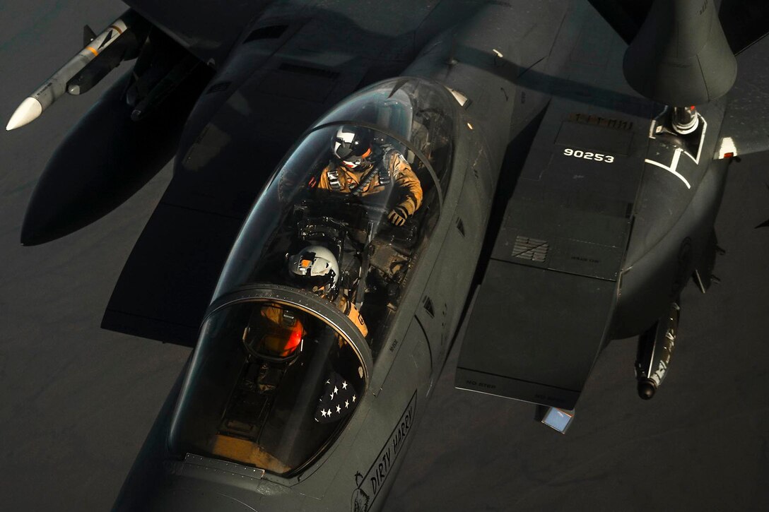 Two airmen are seen from above in the cockpit of a military aircraft in flight.