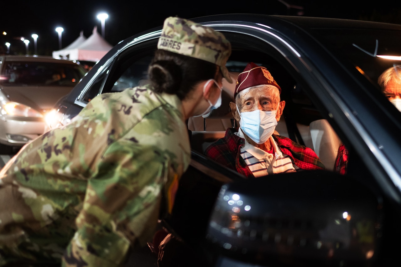A soldier wearing a face mask leans over to a car window to talk to an elderly man wearing a face mask.