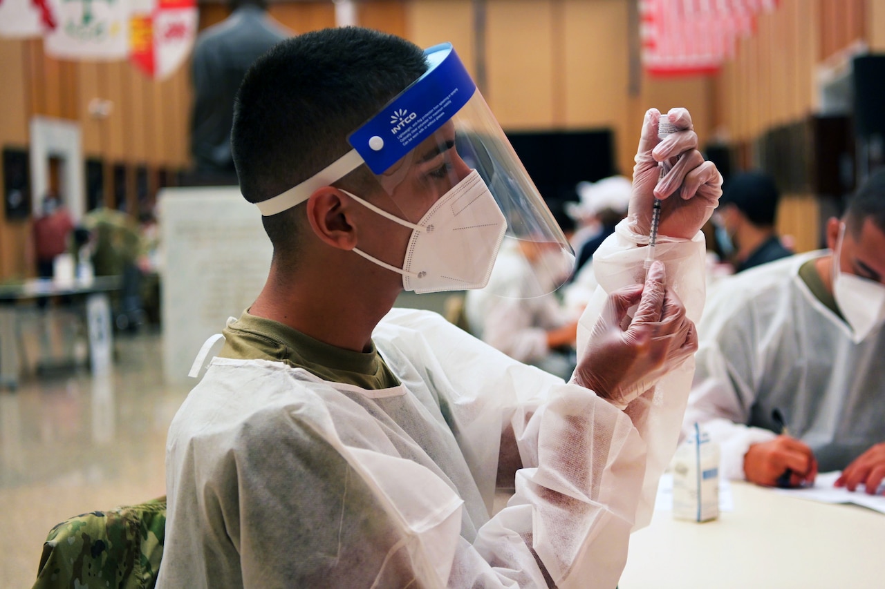 An airman wearing personal protective equipment holds a syringe while inserting the needle into a small bottle.