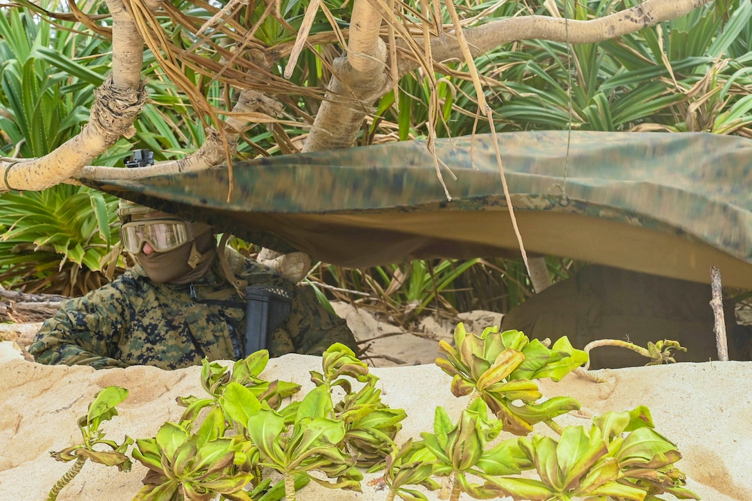 A Marine stands in a sandy hole underneath a tarp.