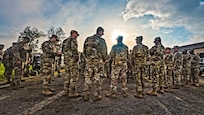 A group of soldiers stand in a group and await orders as the sun rises overhead.