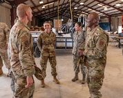 Five soldiers and Airmen wearing fatigues stand in a circle in a staging facility and talk.