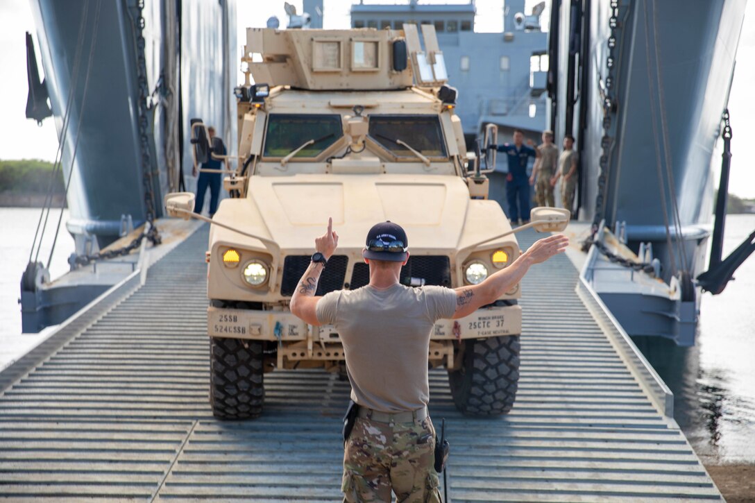 A soldier gives signals to the driver of a military vehicle aboard a water vessel.