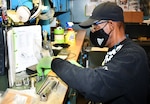 An electrical equipment repairer wearing gloves and a mask is seated at a workbench inventorying parts with reference materials in front of him.