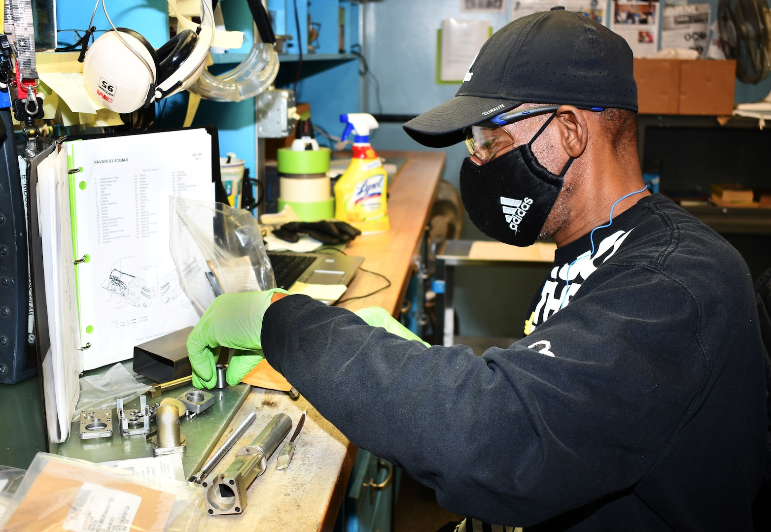 An electrical equipment repairer wearing gloves and a mask is seated at a workbench inventorying parts with reference materials in front of him.