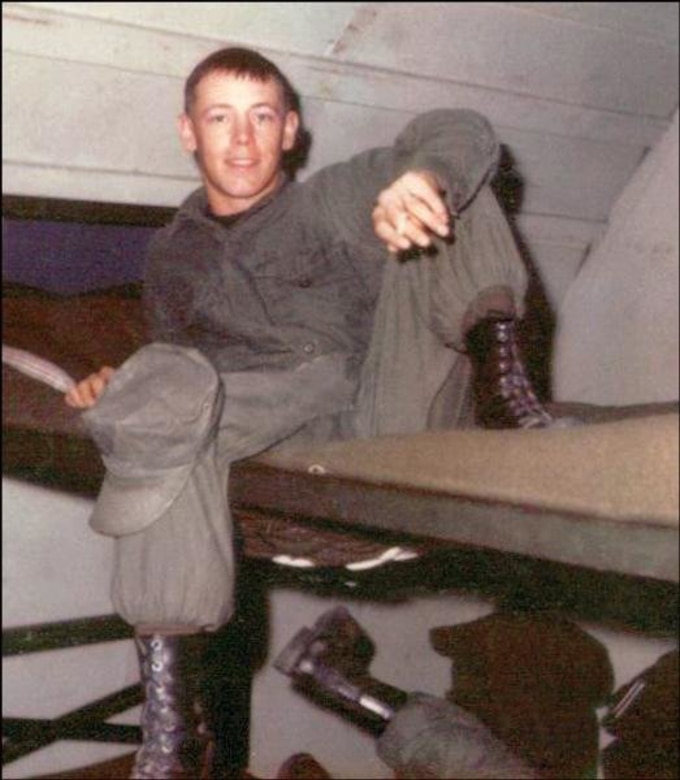 A young man sits with his arm on his knee on the top bunk of a bunk bed.