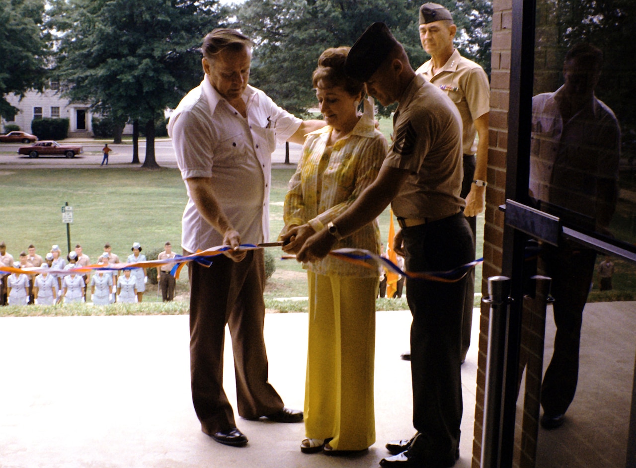Three people stand together to cut a ribbon at the door of a building. Several people watch at the bottom of several steps.