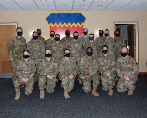 A group of airmen in uniform, six in front row kneeling on one knee, the rest standing behind them. All posing in front of a wall with the 55th Wing shield centered behind them.