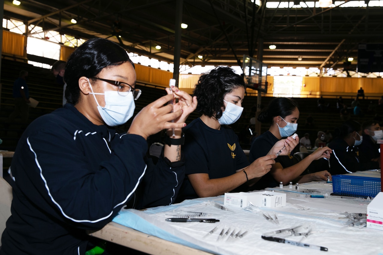 Three women wearing face masks sit at a table using syringes to draw fluid from small bottles.