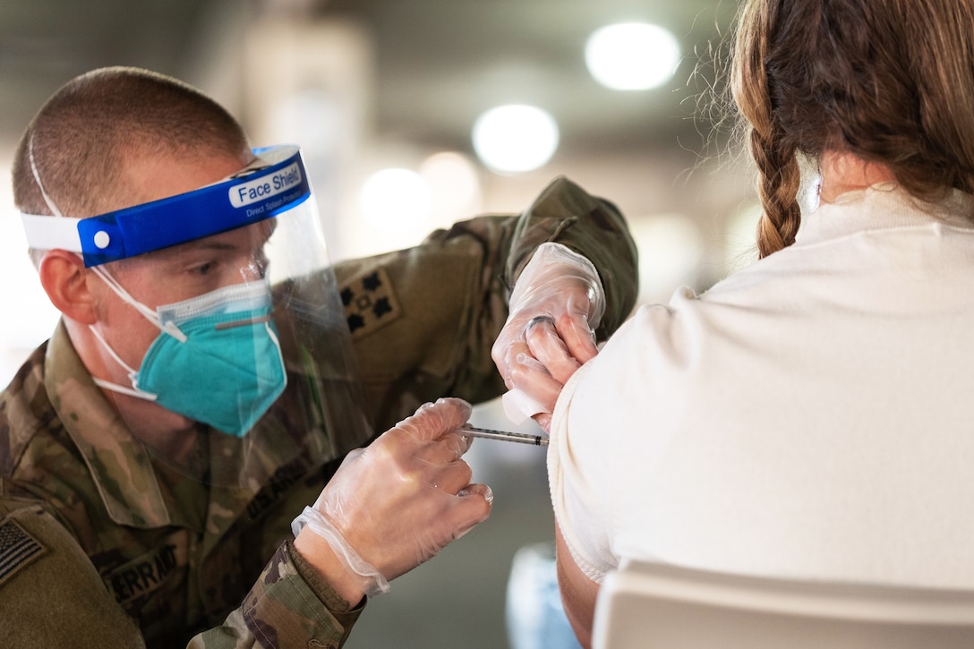 A soldier wearing a face mask and gloves kneels down to give a shot in the arm of a woman sitting in a chair.