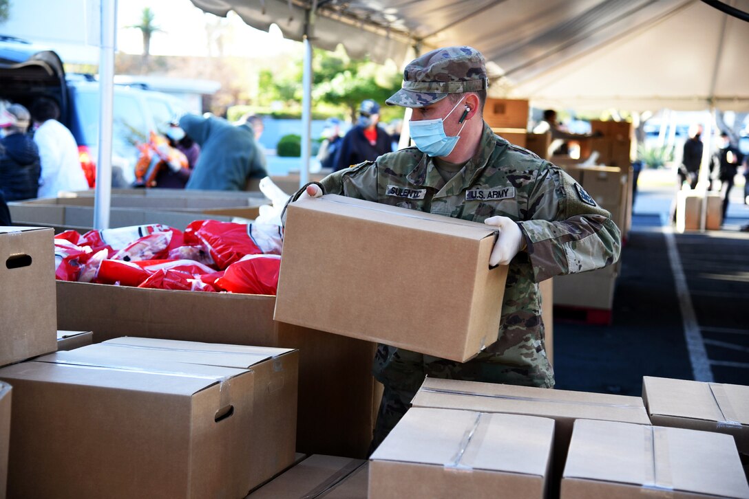 Outside under a tent, a soldier wearing a face mask and gloves picks up a box; other boxes are stacked up in front of him.