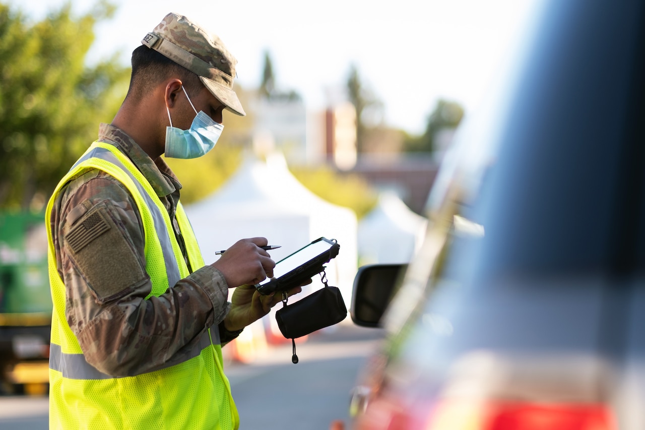 A soldier wearing a face mask looks at an electronic tablet while a motorist waits in a vehicle