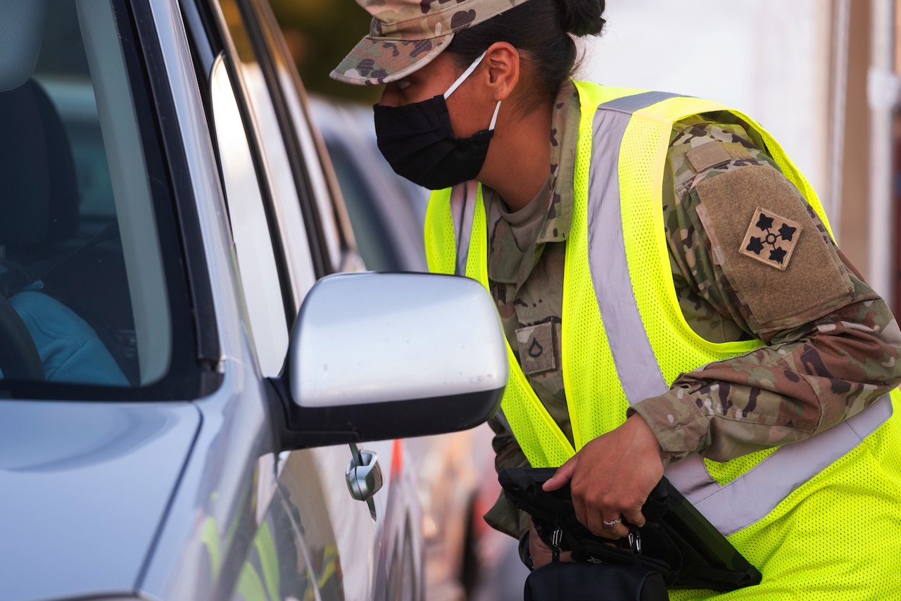 A female soldier wearing a face mask leans into the window of a vehicle.