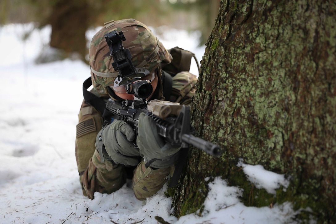 A soldier looks through the scope of a weapon from behind a tree.