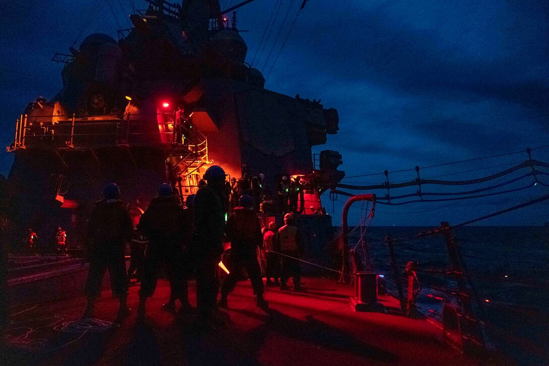 Sailors stand on the deck of a ship at night.