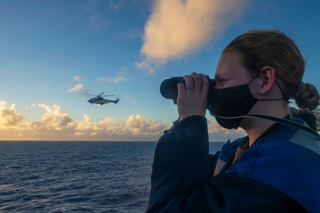 A sailor looks through binoculars from the deck of a ship with an aircraft on the horizon.