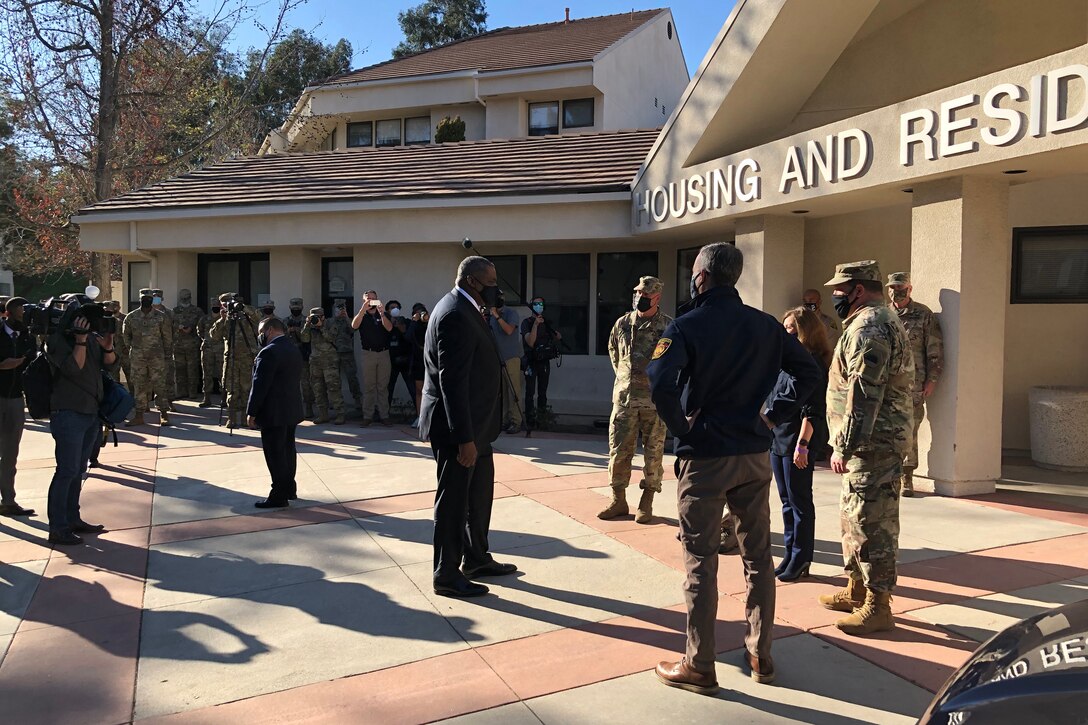 In front of a large building, a man dressed in a business suit stands facing a man dressed in civilian clothes and people in military uniforms.