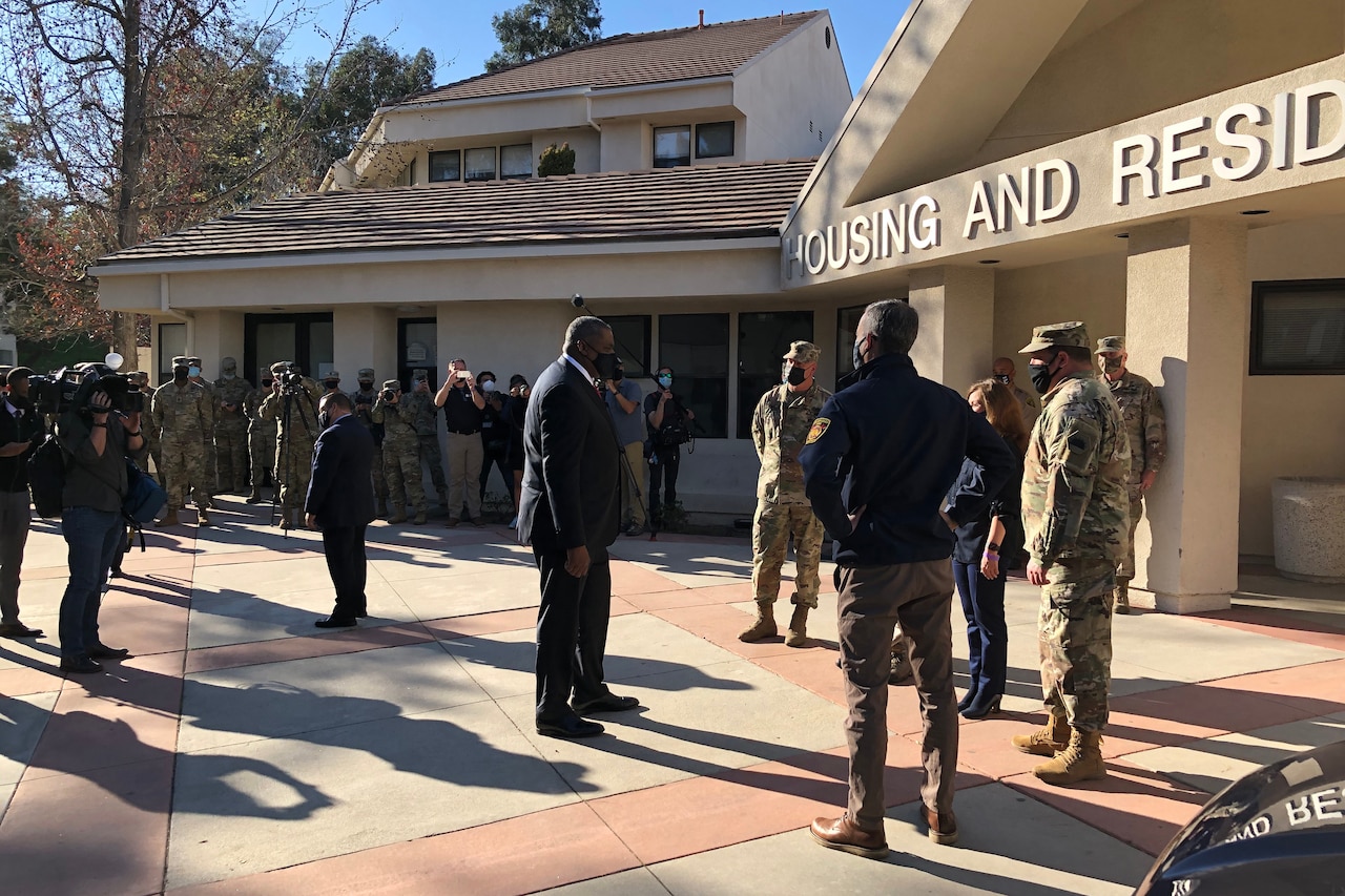 In front of a large building, a man dressed in a business suit stands facing a man dressed in civilian clothes and people in military uniforms.