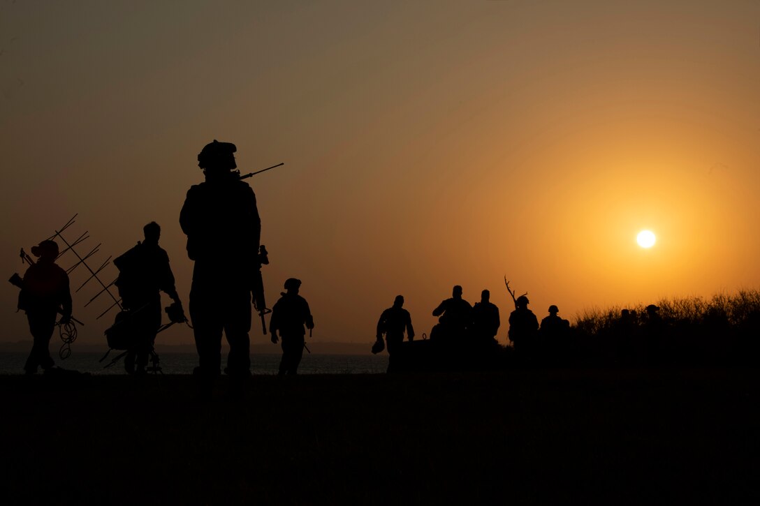 Marines shown in silhouette walk under a sunlit sky.