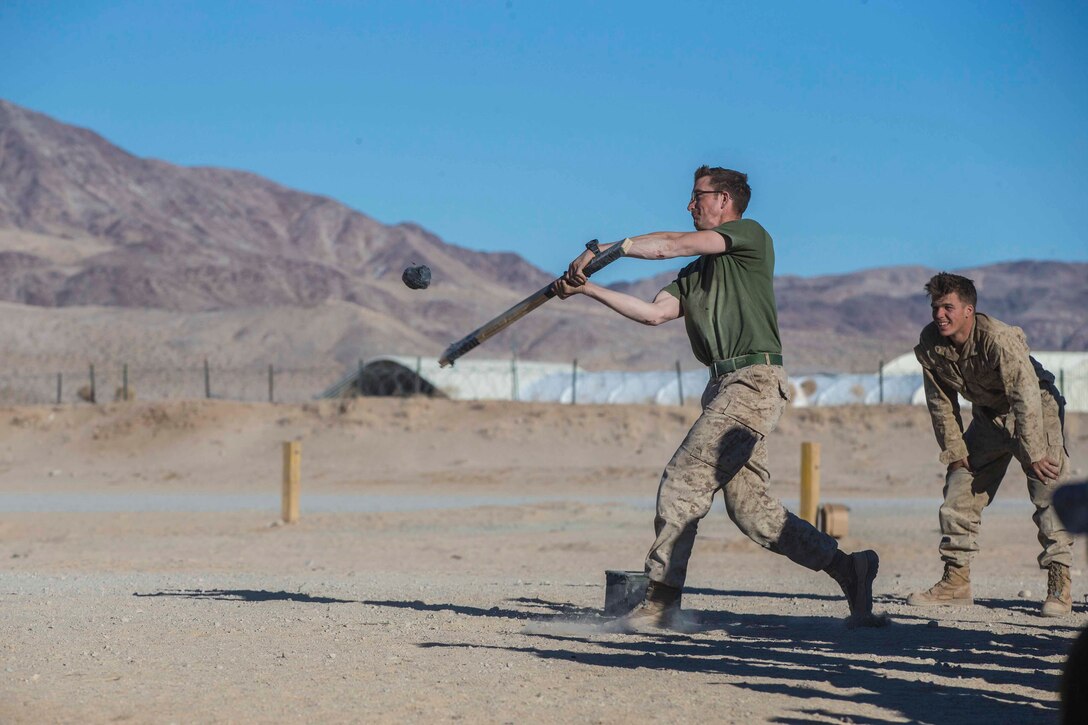 A Marine swings a stick at a ball as another Marine stands behind.