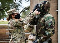An instructor pulls on the vents of a gas mask an Airman in full protective gear is wearing.