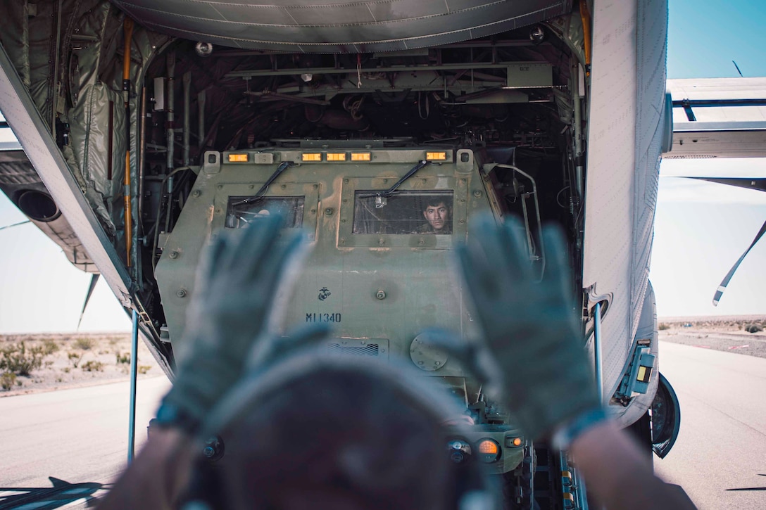 A Marine holds up their hands as another Marine backs a large military vehicle into an aircraft.