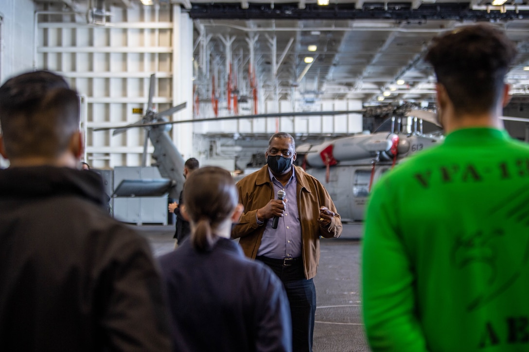 A man wearing a protective face masks speaks to a group of people on an aircraft carrier.