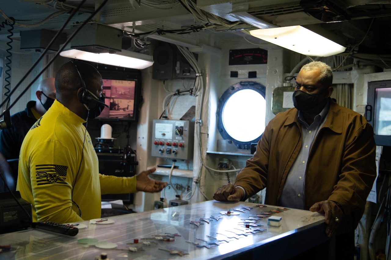 Two men wearing face masks speak in a room on a Navy ship.