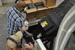 IMAGE: Naval Surface Warfare Center Dahlgren Division Industrial Hygienist Keith Hakanen measures sound levels as Information Security Specialist Jason Gray places a hard drive on the GigaBiter’s conveyer belt for its ultimate destruction.