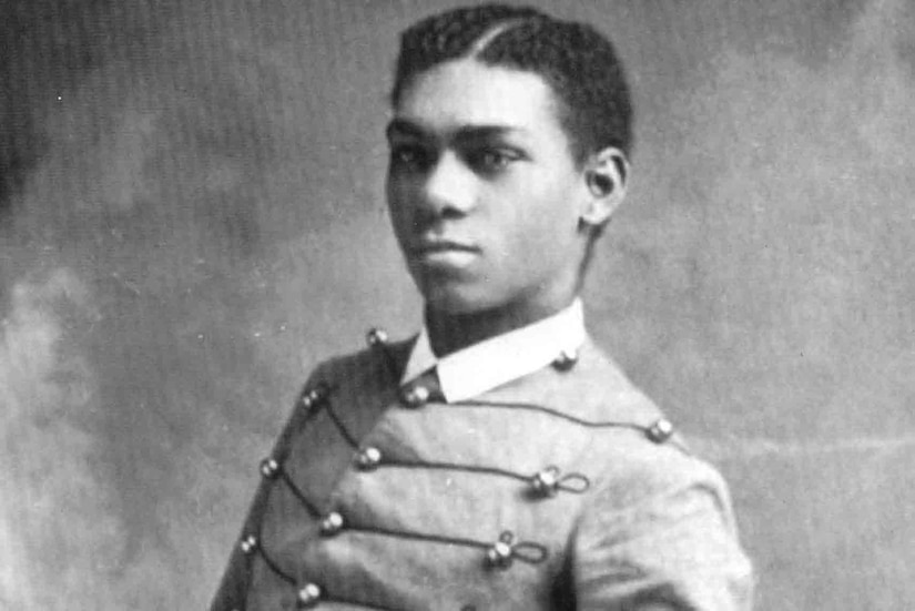 A historic photo shows a young man standing with his arm on the back of a chair.