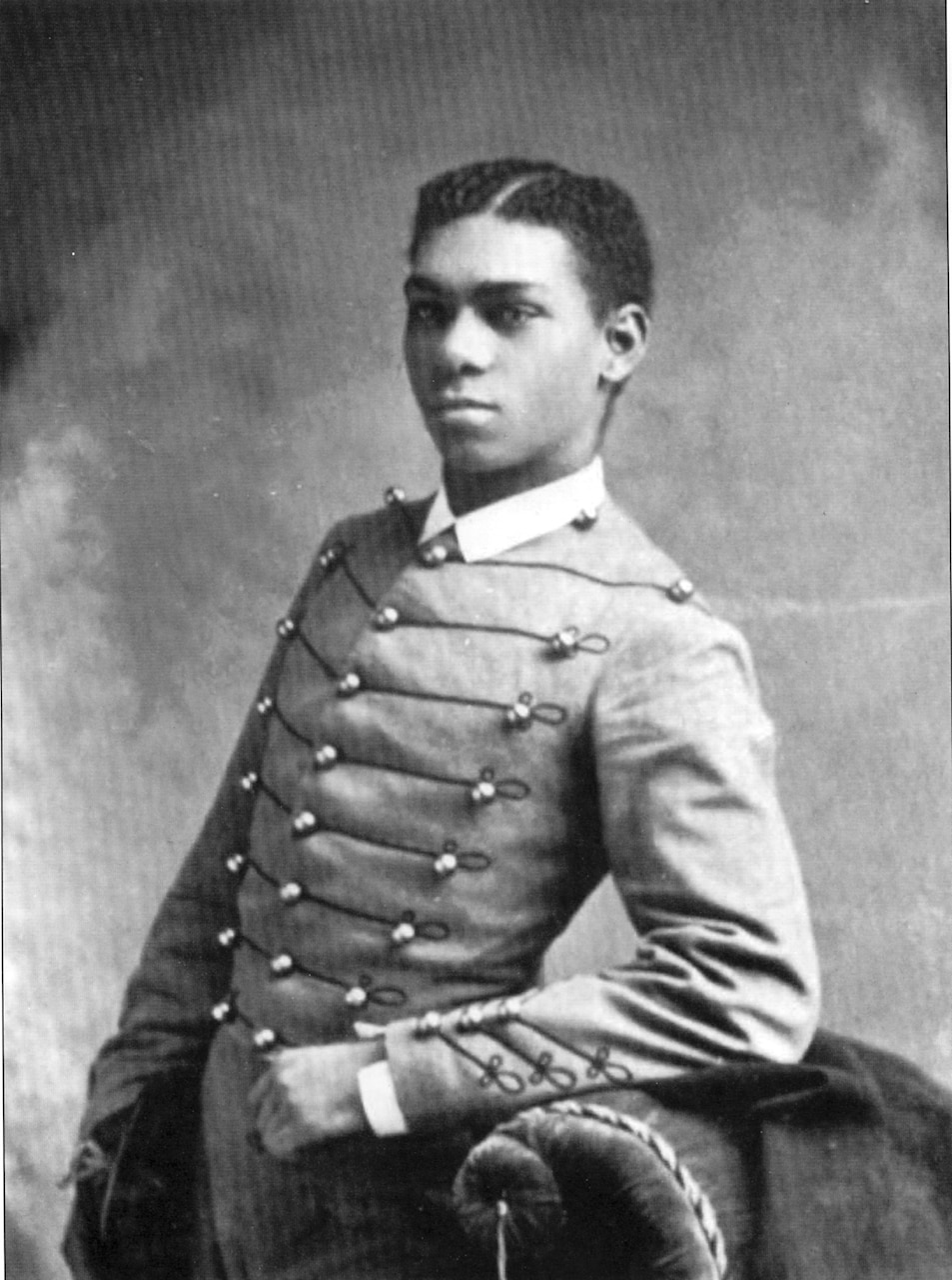 A historic photo shows a young man standing with his arm on the back of a chair.