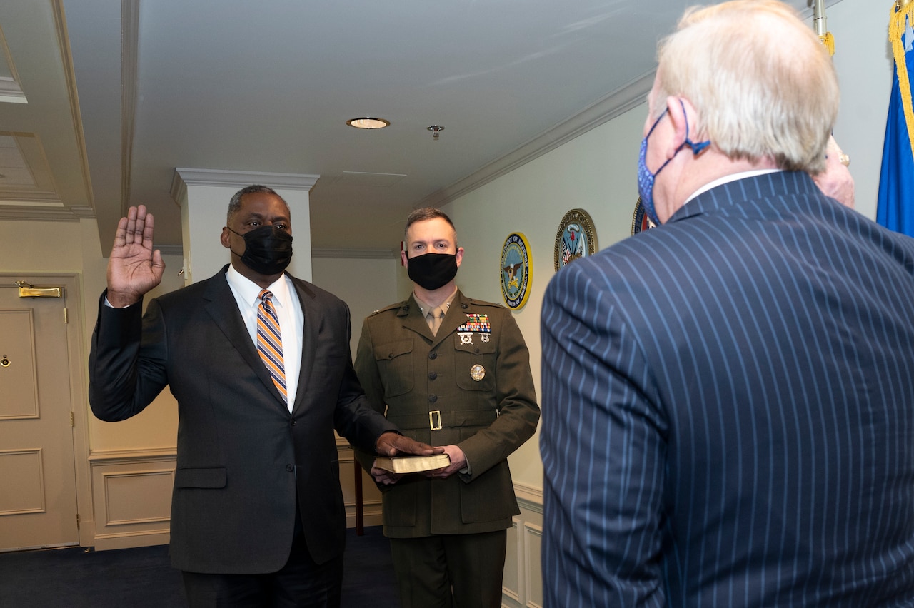 A man dressed in a business suit stands in an office with his left hand on the Bible and his right hand raised. A man wearing a military uniform holds the Bible; another man with his back to the camera is in the foreground. All three men wear face masks.