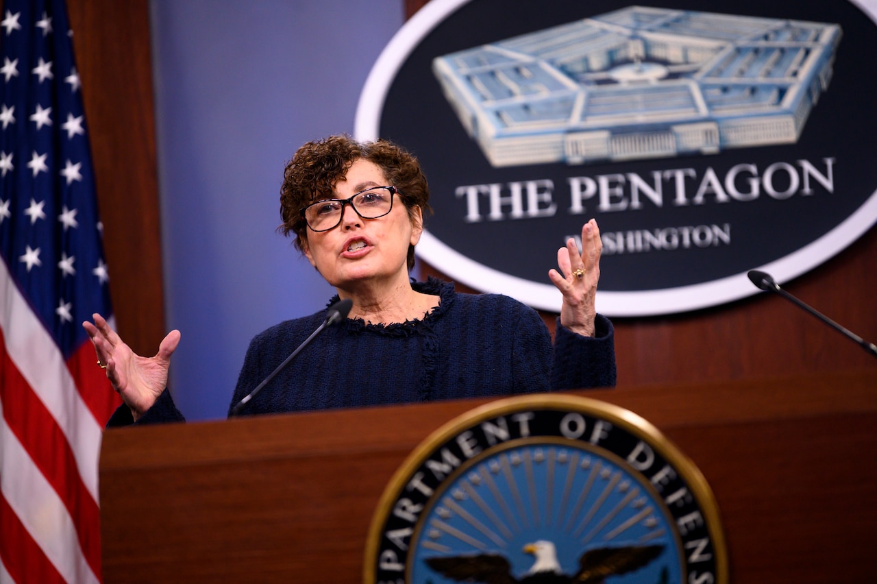 A woman stands behind a lectern. In the rear, a sign on the wall reads "The Pentagon - Washington."