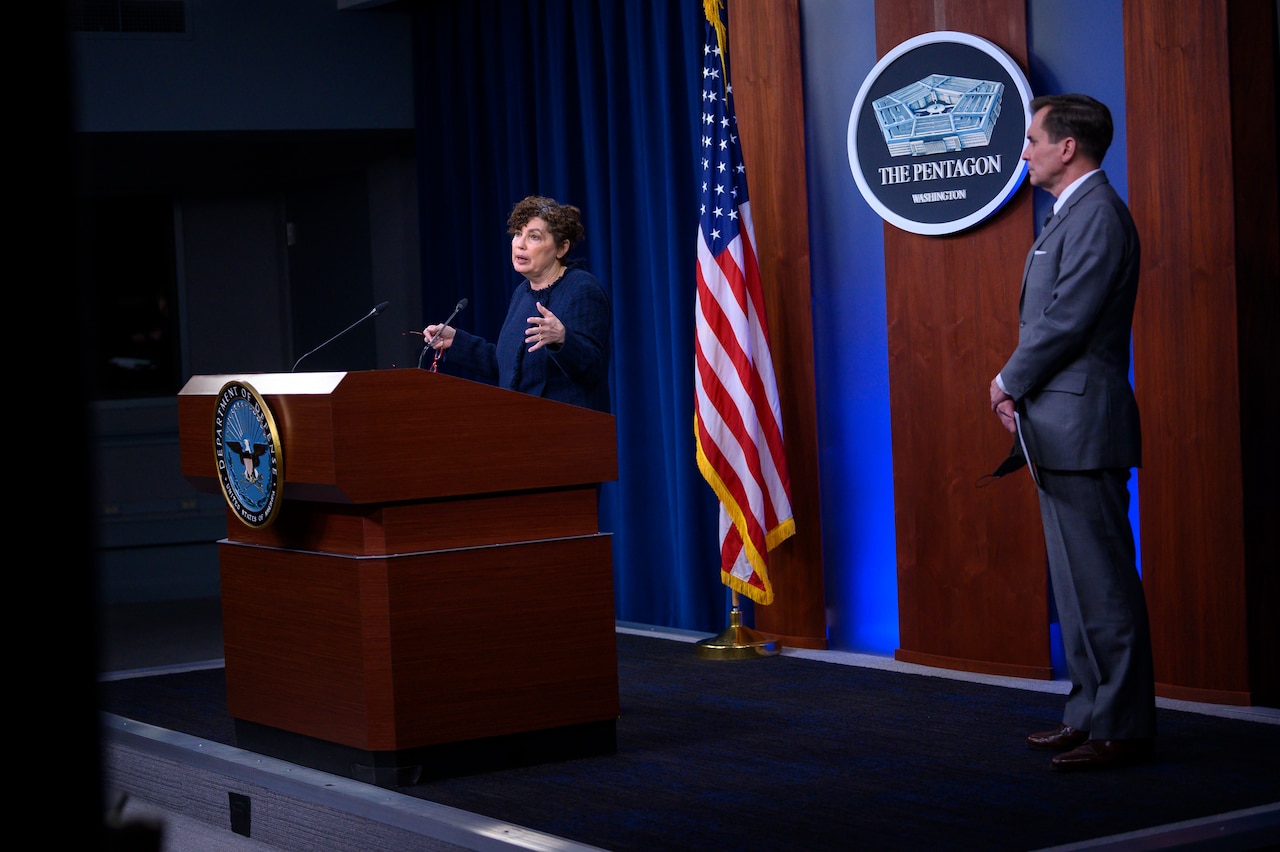 A woman stands behind a lectern. Nearby, a man stands with his arms folded in front of him.