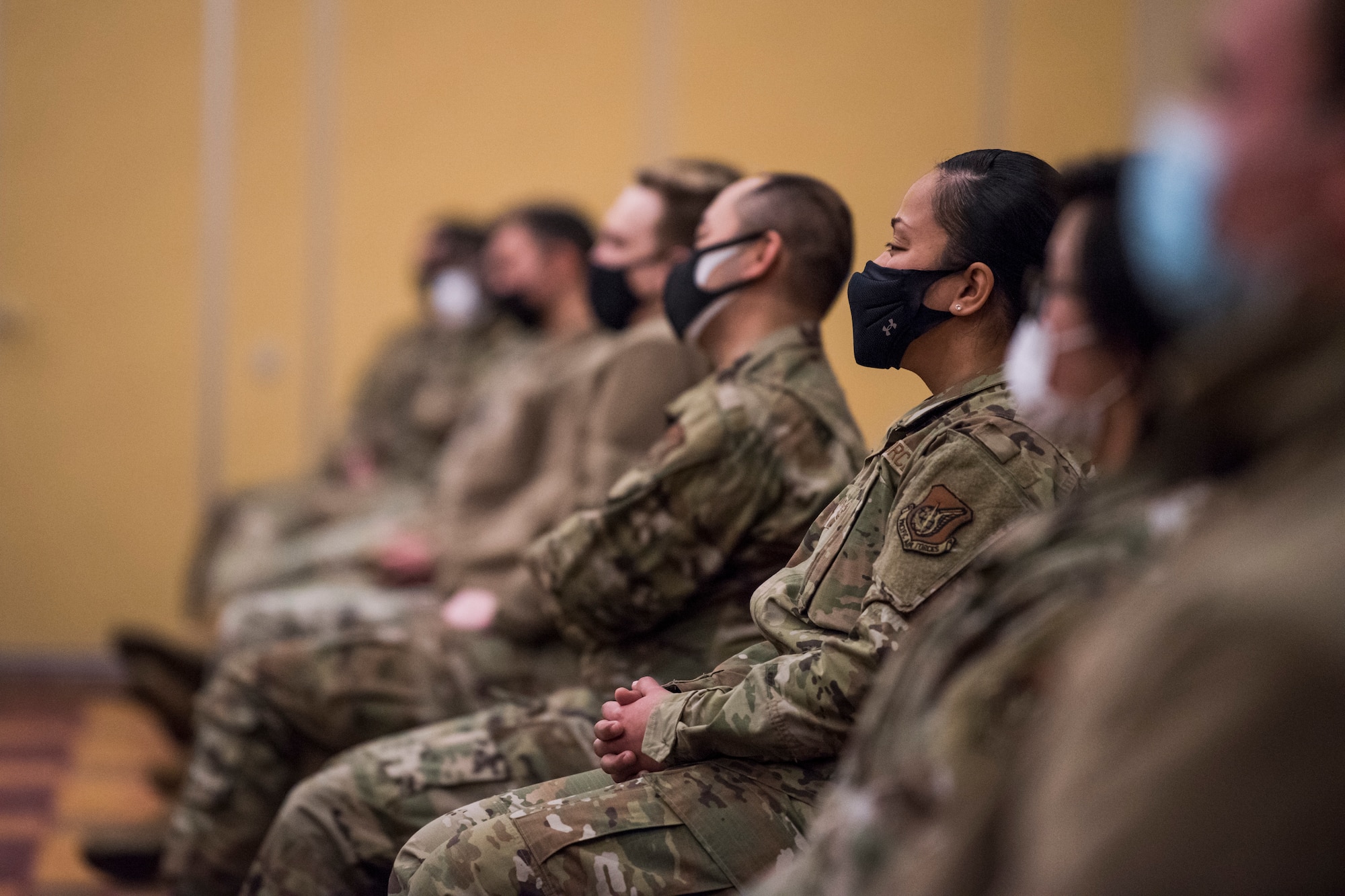 Military Members with masks sit in a room listening.