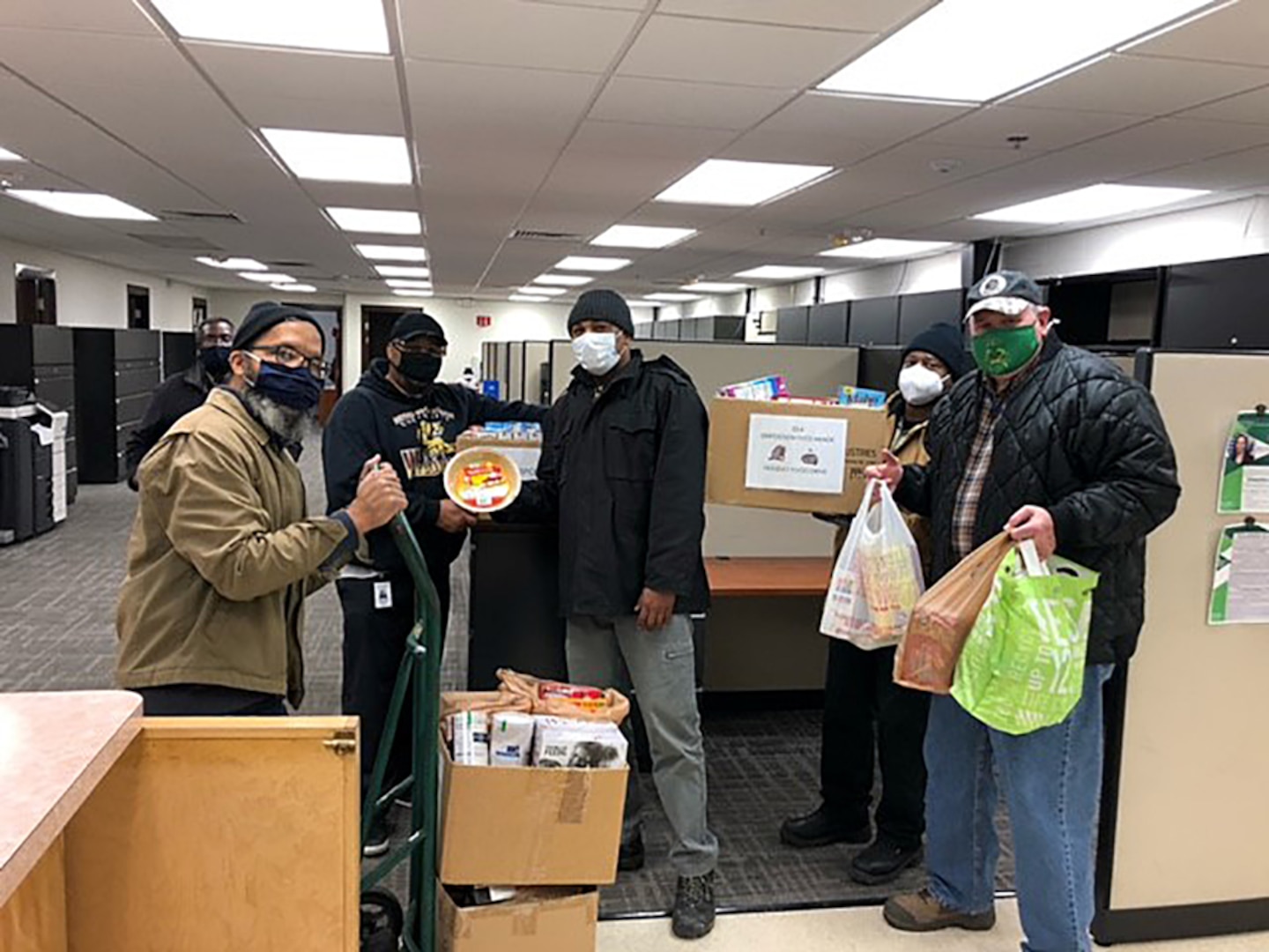Masked men stand in an office space with boxes and bags of food items for donation.