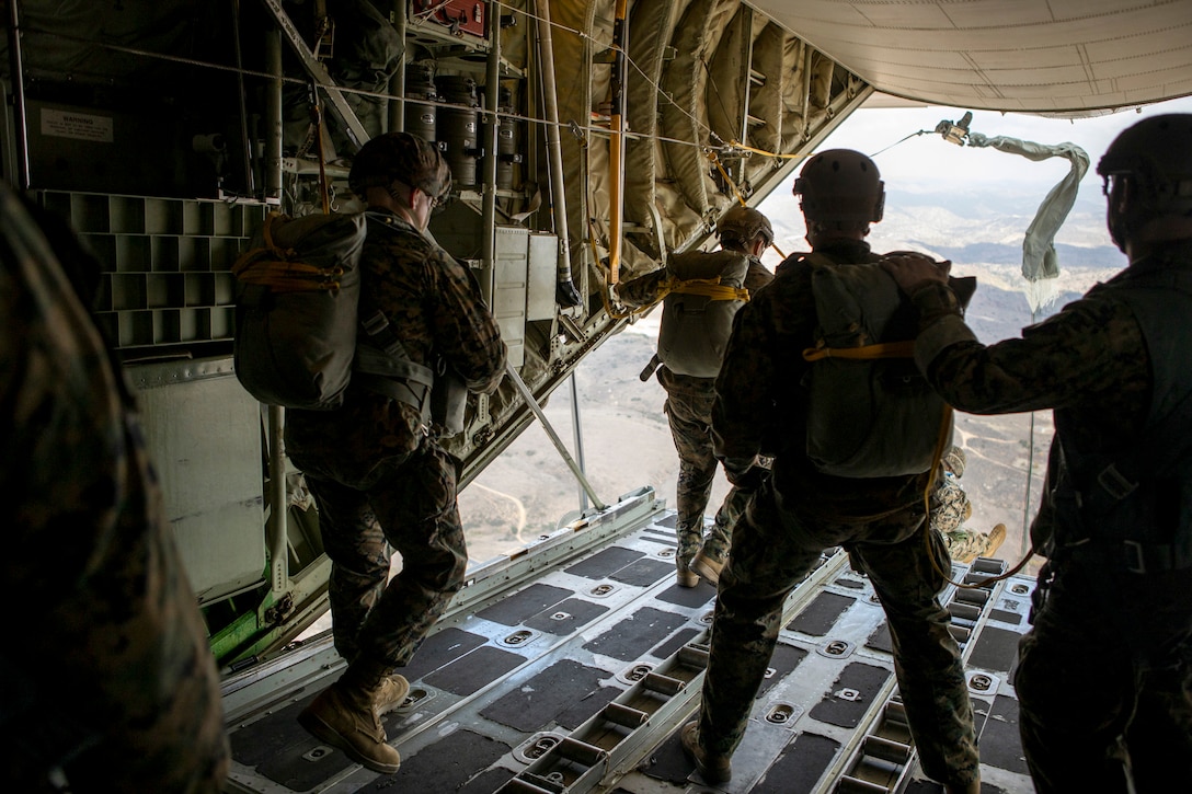 Several Marines stand at the back of an open aircraft in flight as one jumps.