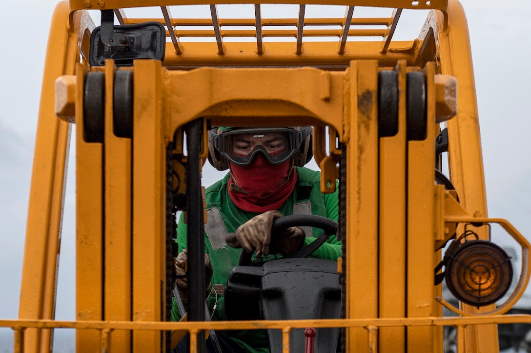 A sailor in green drives a yellow forklift.